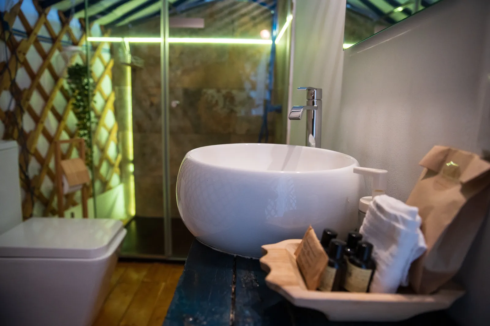 El baño del refugio: lavabo redondo de porcelana sobre una repisa de madera, cabina de ducha acristalada al fondo y toallas y jabones preparados en una bandeja