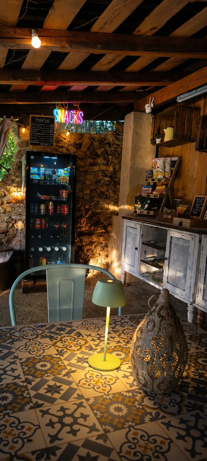 El bar honesto de la finca de noche: una nevera con bebidas, un cartel de neón que dice «Snacks», la lista de precios en una pizarra y una mesa con azulejos y un farol encendido