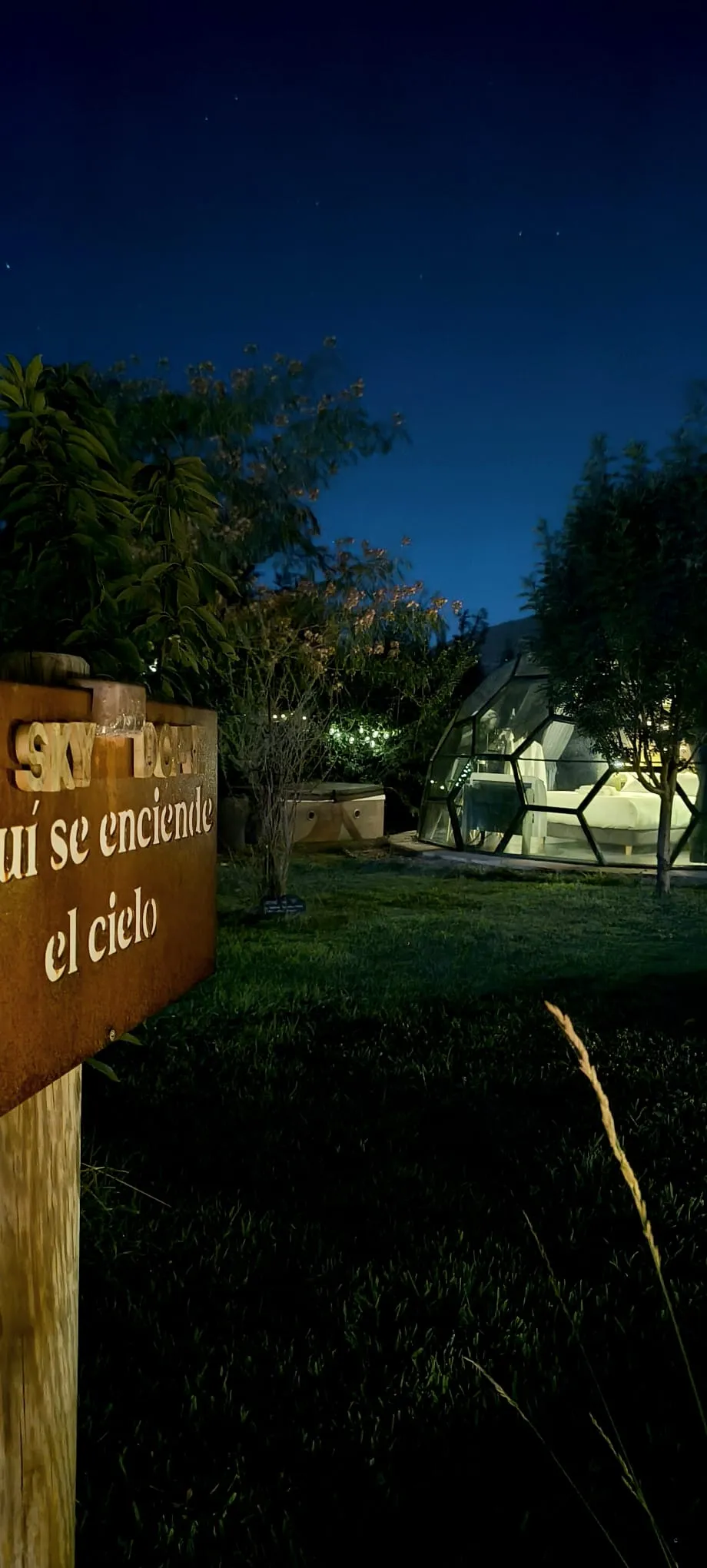 El cartel de acero oxidado que dice «Aquí se enciende el cielo» sobre su poste de madera, de noche, con el domo geodésico encendido por dentro y el jacuzzi al fondo, sobre el césped