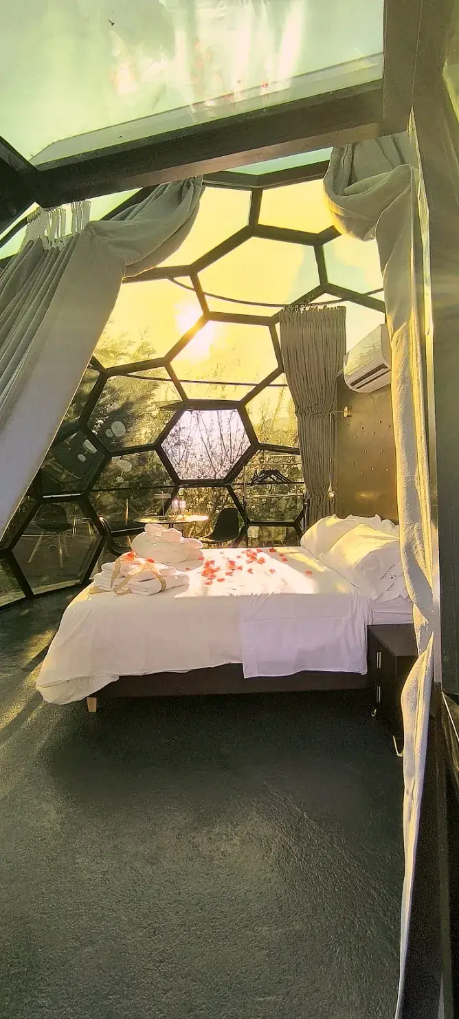 La cama del Sky Dome preparada con pétalos de rosa y toallas dobladas sobre la colcha blanca, bajo la bóveda hexagonal de cristal, con la luz del atardecer entrando de frente