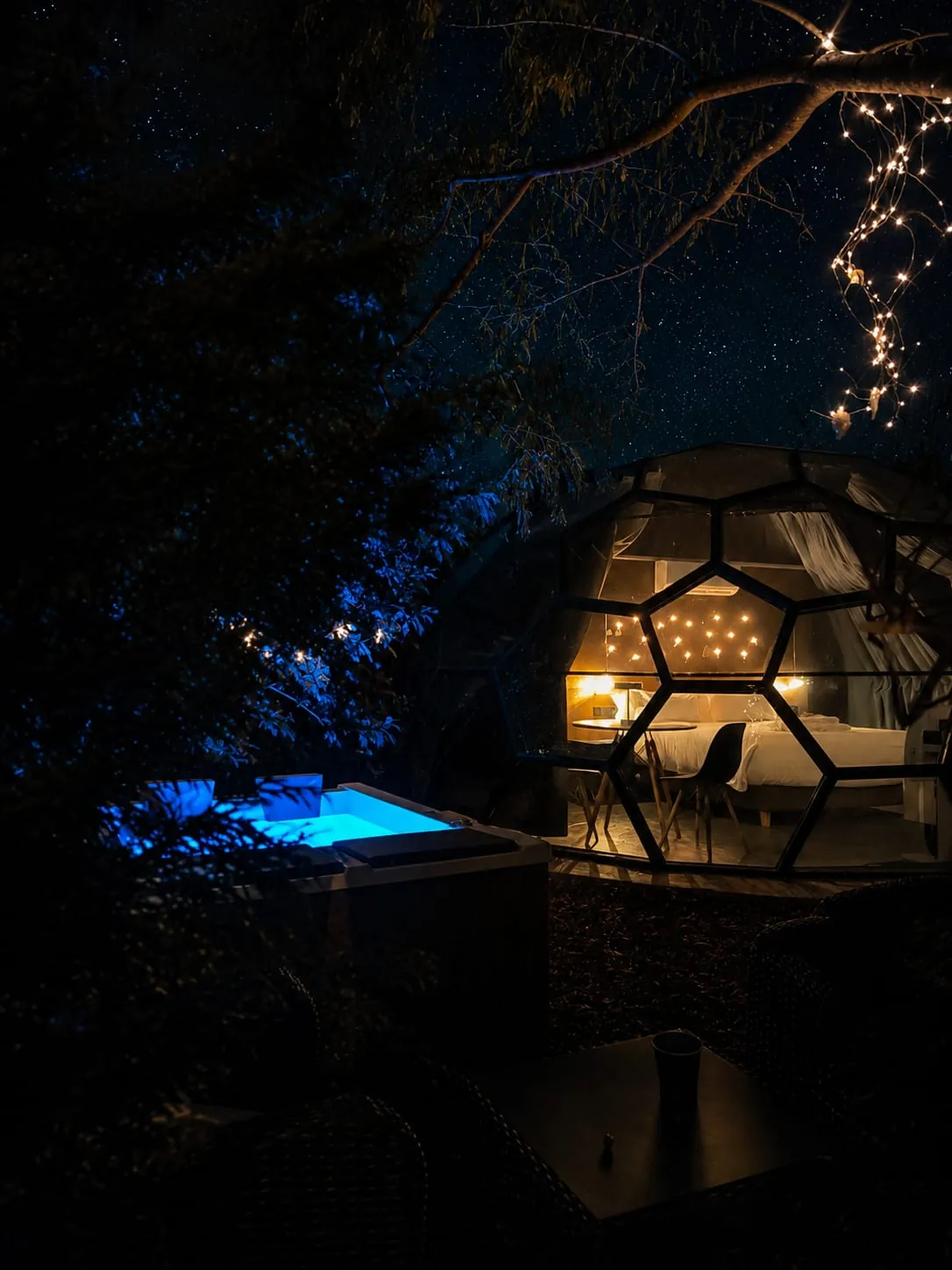 El Sky Dome iluminado por dentro visto de noche entre los árboles: por el cristal se ven la cama y las lucecitas del cabecero, con el jacuzzi encendido en azul a la izquierda y una guirnalda de luces colgando de una rama