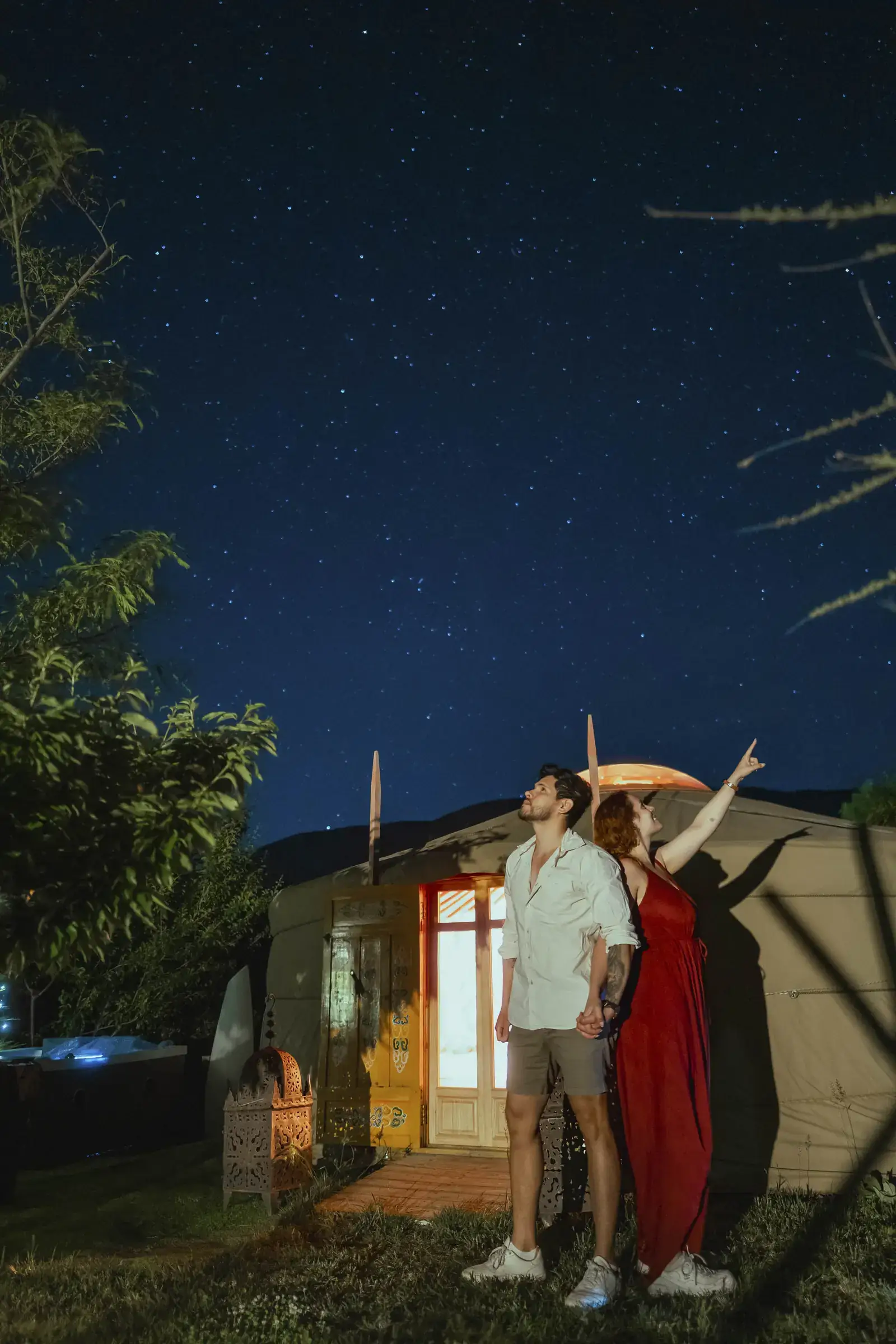 Una pareja de noche delante de una yurta encendida por dentro: ella señala una estrella con el brazo levantado y él mira hacia arriba, bajo un cielo lleno de estrellas