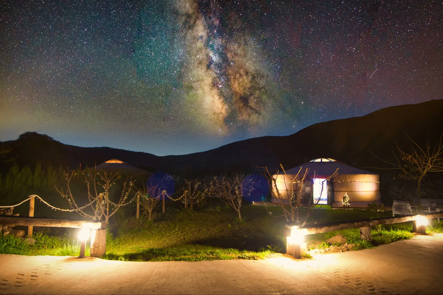 La Vía Láctea sobre la finca de noche, con dos yurtas iluminadas por dentro entre los árboles y la sierra recortada en negro al fondo
