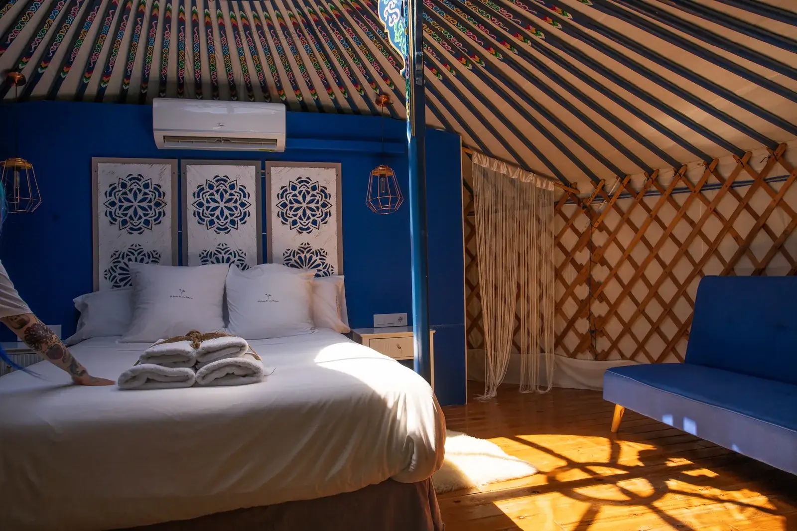 La corona de la Yurta Cielo pintada a mano, vista desde la cama: franjas de colores que convergen hacia el centro, con la celosía de madera y el sofá azul al fondo, iluminados por el sol de la tarde