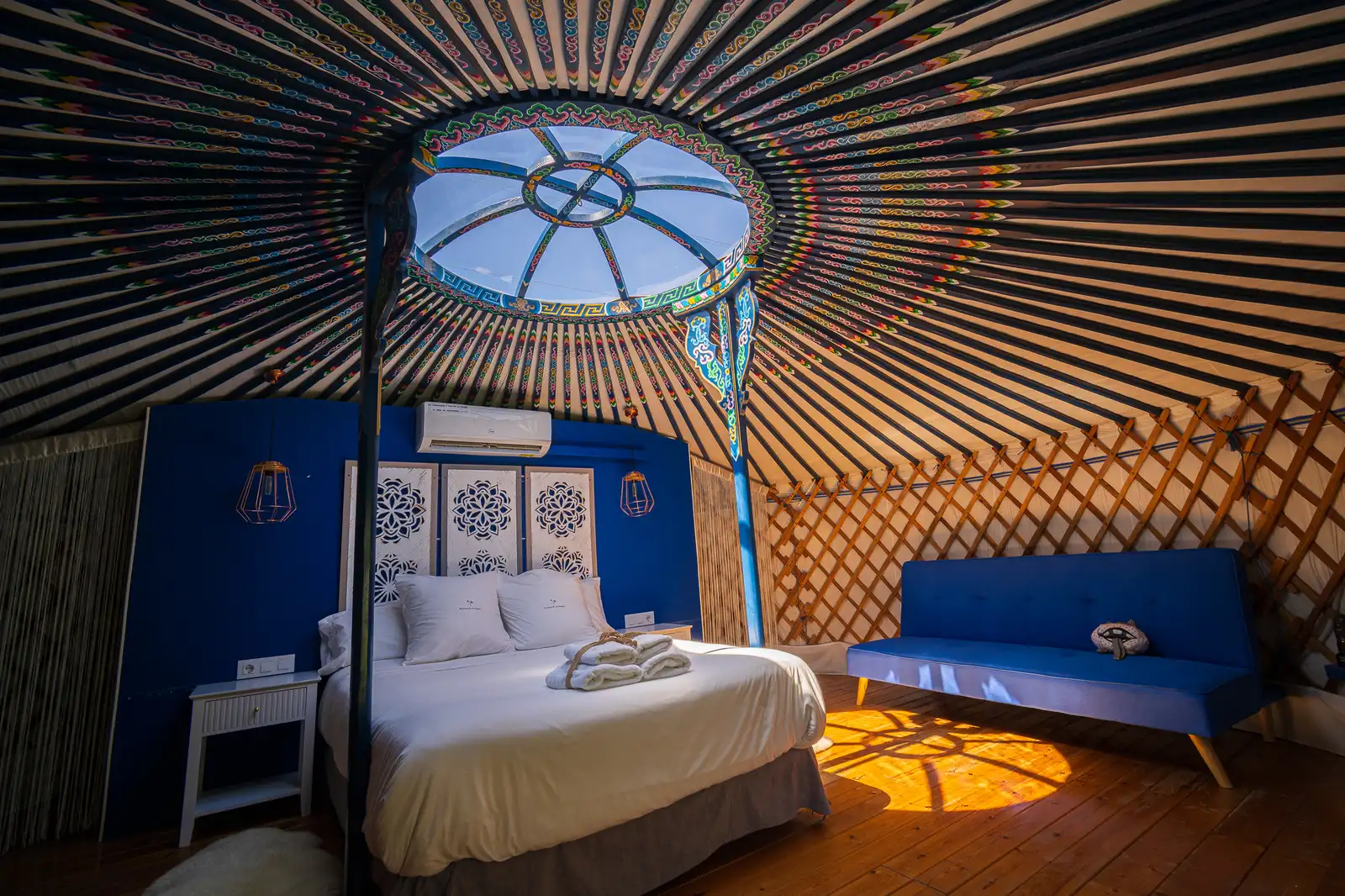 El interior completo de la Yurta Cielo desde los pies de la cama: la corona pintada, el óculo, el sofá azul y la celosía de madera con la luz de la tarde entrando